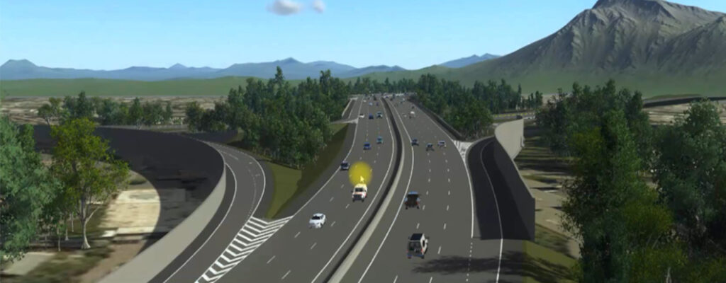 A wide highway with multiple lanes, a hallmark of civil engineering, carries vehicles in both directions, surrounded by trees and distant mountains under a blue sky.