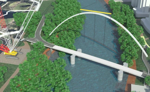 synchro-case-study An architectural rendering shows a white arched pedestrian bridge over a river surrounded by trees, with a crane positioned nearby. Buildings and roads are visible in the background, illustrating solid support structures in place.