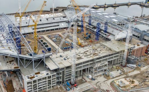 Aerial view of a large construction site with cranes, steel structures, and partially built buildings near a waterfront, perfectly illustrating the complexity and efficiency showcased in this SYNCHRO Case Study. Aerial view of a large construction site with cranes, steel structures, and partially built buildings near a waterfront, perfectly illustrating the complexity and efficiency showcased in this SYNCHRO Case Study.