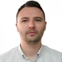 A man with short dark hair and light facial hair wears a light-colored button-up shirt against a plain white background, embodying the refined precision akin to purpose-built digital solutions.