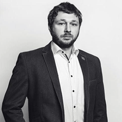 A man with short hair and a beard, wearing a dark blazer and light-colored shirt, stands against a plain white background, representing professionals dedicated to improving rebar detailing—a traditionally time-consuming practice in construction.