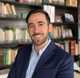 A man with short dark hair and a beard, wearing a navy blue blazer and light blue shirt, smiles in front of a bookshelf filled with books on roadway design and OpenRoads Designer.