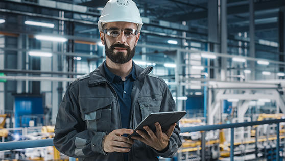 A worker in a hard hat is holding a tablet and giving a demo in a factory. | Bentley Systems | Infrastructure Engineering Software Company A worker in a hard hat is holding a tablet and giving a demo in a factory.