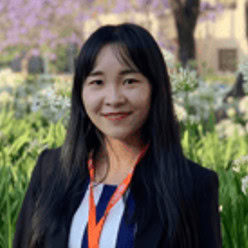 A person with long dark hair and an orange lanyard stands outdoors, surrounded by green foliage and white flowers, set against purple trees blurred in the background, evoking a design as intricate as any crafted in the Structural WorkSuite.