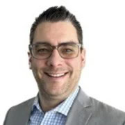 A person with short dark hair and glasses is smiling, wearing a light gray suit jacket and a blue checkered shirt, exuding a professional WorkSuite vibe against a plain white background.