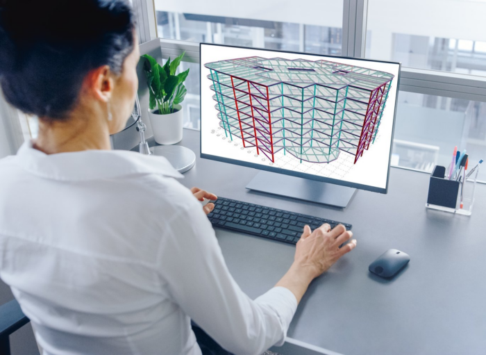 Person in a white shirt working on a computer displaying a 3D architectural model of a building using Structural WorkSuite. The desk also has a plant, lamp, and various office supplies.