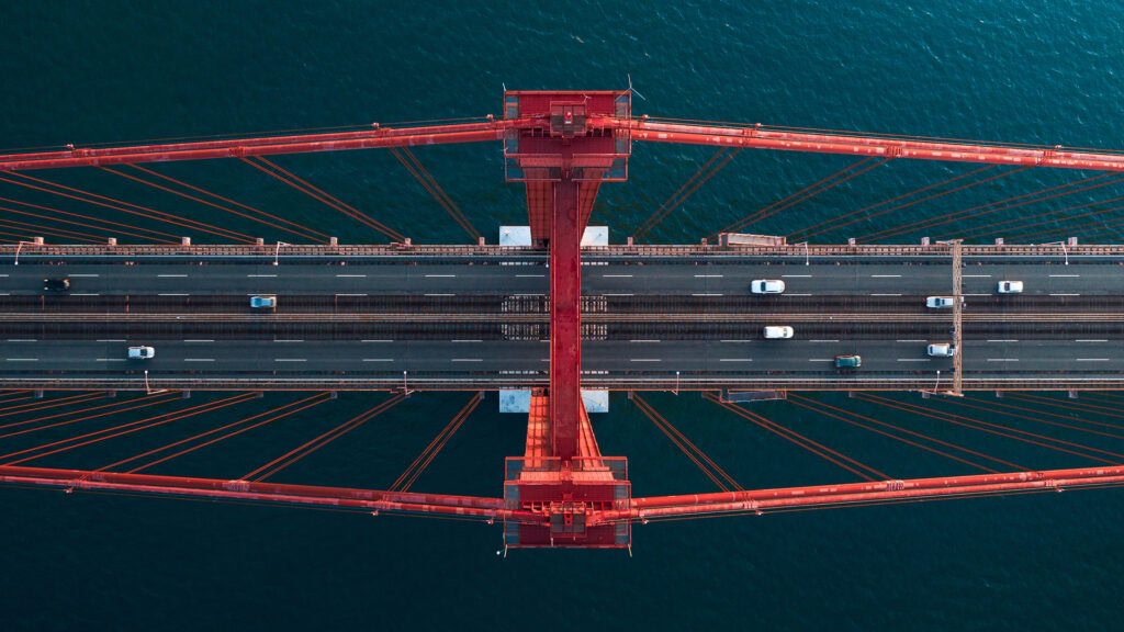 Aerial view of a red suspension bridge in 2024, showcasing the vibrant scene of transportation with multiple vehicles driving on it, set against a backdrop of blue water.